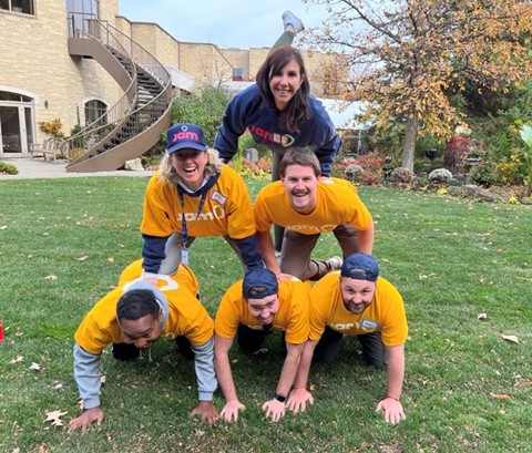Kristi Herold and 5 of her work friends (JAM teammates) form a human pyramid. They are wearing yellow JAM t-shirts. 